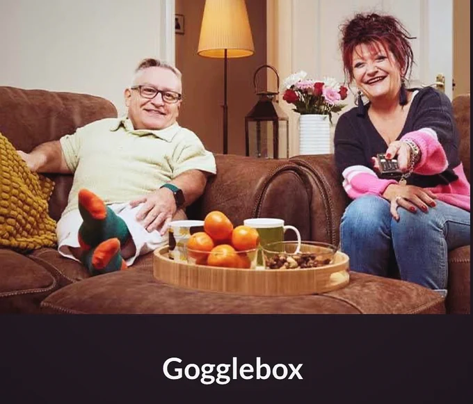 Simon on the left in a pale green top and Jane on the right in a blue and pink jumper. Both sat on a sofa and Jane’s holding a tv remote.