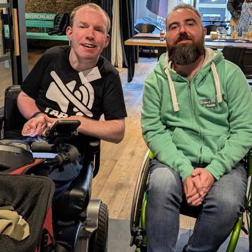 Image of Lost Voice Guy, a clean-shaven white man with short hair who is wearing a black T-shirt with a logo reading Tap To Unmute, he sits beside Joe, a white man with a short beard. Joe wears a green Euan’s Guide hoodie.  Lost Voice Guy sits in his scooter, and Joe in his wheelchair
