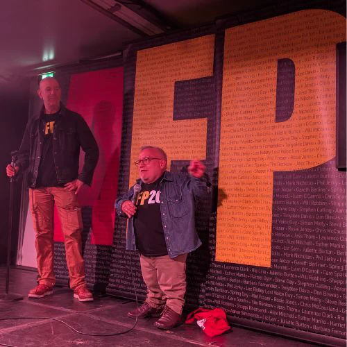 Image of  Simon Minty and Steve Best in front of AFP signage. Simon is a white man with short grey hair and glasses, wearing a blue jean jacket and a black AFP20 branded Tee

Steve, a white man with a shaved head, wears the same tee and a dark jacket and is looking at Simon

