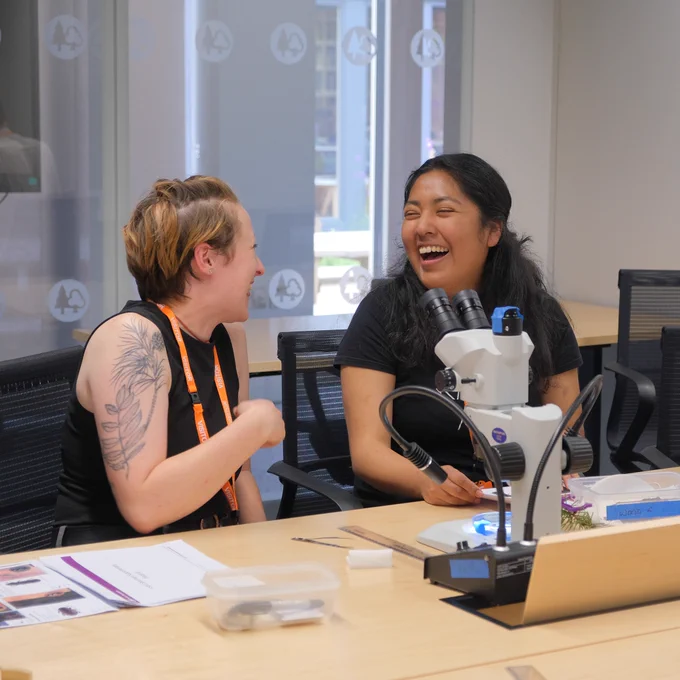 Two people sitting at desk and laughing. Microscope in foreground. 