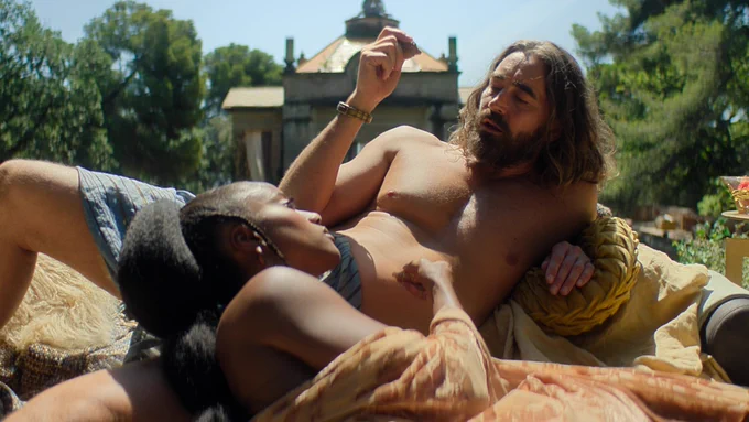 Lee Pace as Brother Day lounges shirtless on a daybed outdoors while Yootha Wong-Loi-Sing as Song lays facing the opposite way with him.