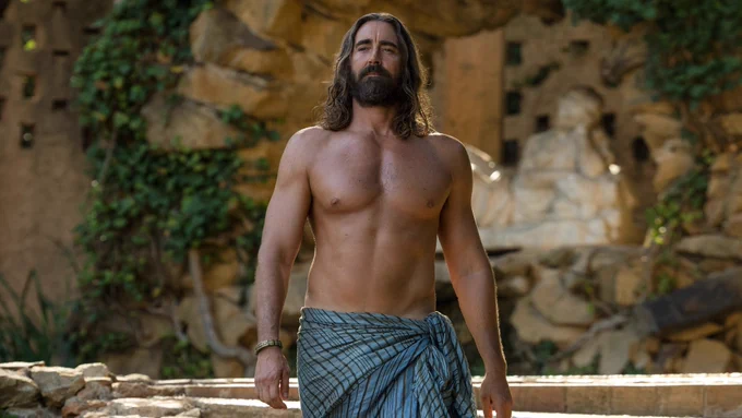 Lee Pace as Brother Day stands shirtless confidently with blue linen wrapped around his waist.