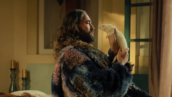 Lee Pace as Brother Day sits in a large fur coat holding a white ferret up to his face in profile view.