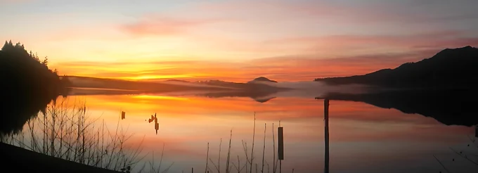 A panoramic view of a calm lake at sunrise, with the sky ablaze in vivid gold, orange, and soft pink hues. The still water mirrors the colors almost perfectly, creating a symmetrical reflection of the silhouetted mountains and sparse shoreline vegetation. A few weathered wooden posts rise from the water, adding depth and texture, while a faint mist lingers near the distant horizon.