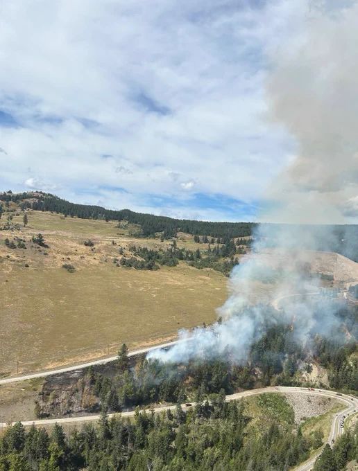 Smoke billows from a treed area between two roadways under a partially cloudy sky