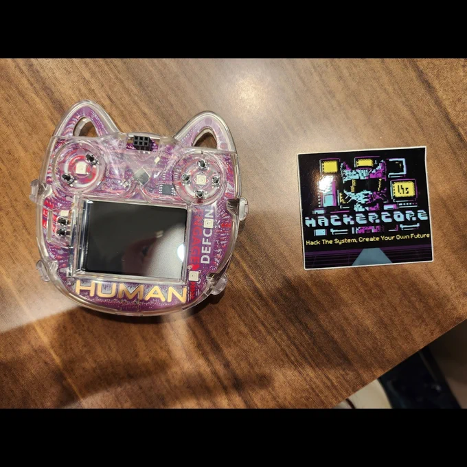 The aforementioned Hackercore sticker sits on a table next to a cat-head-shaped badge reader device. 