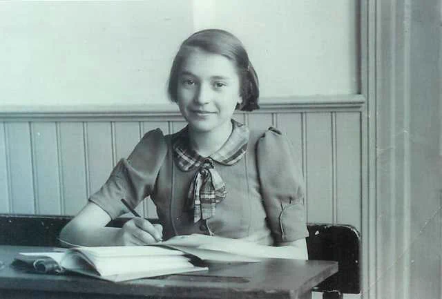 Photograph of teenage girl sitting at a desk, smiling slightly while writing in a notebook.
