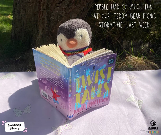 A photograph of a toy penguin (Pebble, the Surrey Libraries Rhymetime mascot) reading a Julia Donaldson book on a picnic blanket under a tree with text. ‘Pebble had so much fun at our 
‘Teddy Bear Picnic Storytime’ last week!‘ The Godalming Library and Surrey Libraries logos are in the corners of the image. 