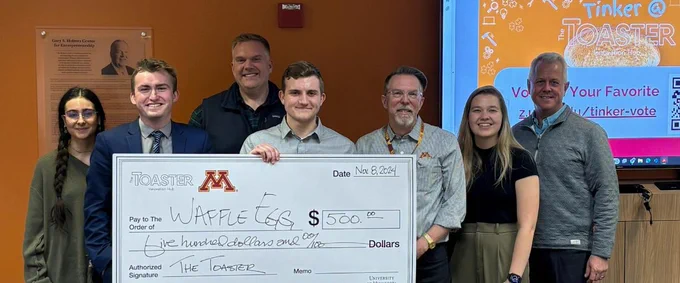 Group of six individuals smiling and holding a large ceremonial check from The Toaster for $500 written to The Waffle Eggs, at the University of Minnesota.