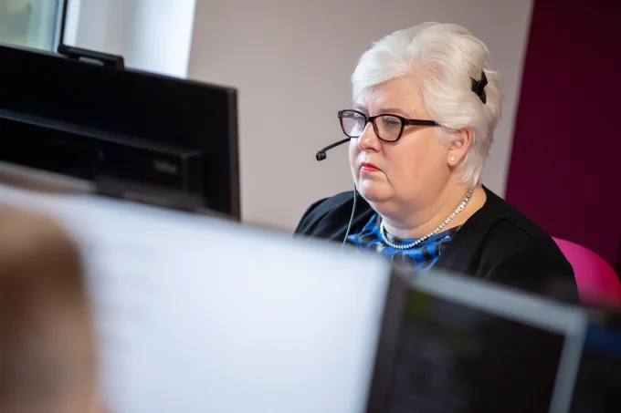 Woman with white hair wearing a headset and glasses working at a computer in an office setting.