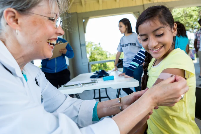Smiling health care worker places a bandage on a young girl’s arm at an outdoor clinic.