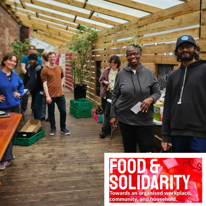 Group of Food & Solidarity volunteers standing and smiling under a wooden-roofed shelter with string lights. Some are gathered near a table with supplies, while others stand along the side with green crates and food items. The setting appears friendly and collaborative, with people dressed casually and chatting.