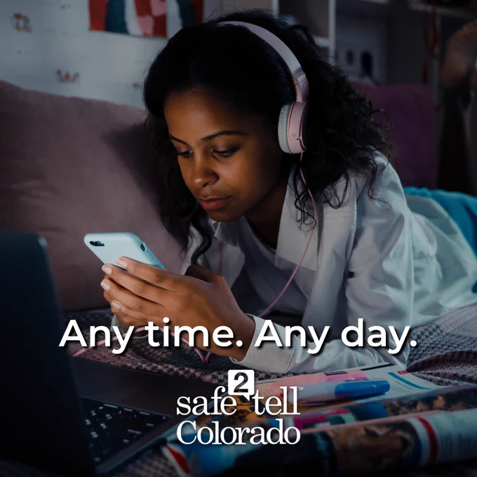 Person using a smartphone while wearing headphones, surrounded by books and notepads, with the text "Any time. Any day. Safe2Tell Colorado" displayed.