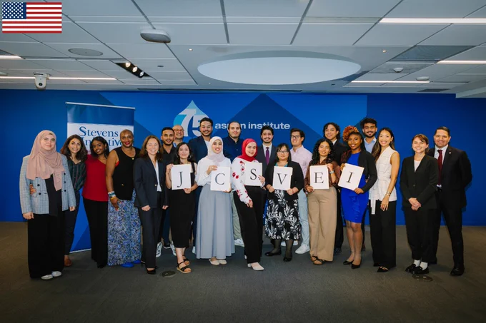Alumni Fellows and stakeholders pose, standing in front of a blue backdrop with the Aspen Institute logo. Several Fellows hold individual letters that together spell “JCSVEI.” 