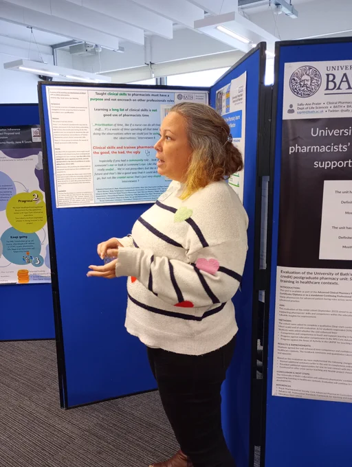 A woman standing in front of a research poster at an academic conference. She is wearing a white jumper with navy stripes and talking to someone not visible in the picture.