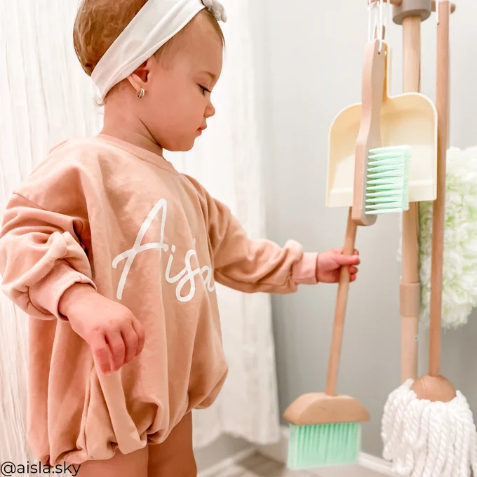 A toddler in a peach-colored shirt with the name "Aisla" printed on the back holding a brush, standing next to a wooden rack with cleaning tools.