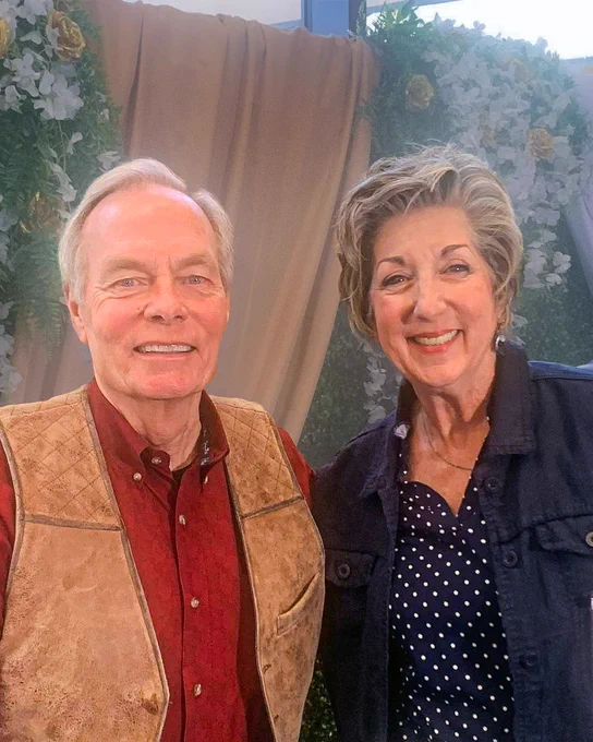 D20 school board candidate Cynthia Halverson smiling with Andrew Wommack, founder of Charis Bible College, at an event decorated with floral backdrops. Halverson is wearing a Charis Bible College badge.