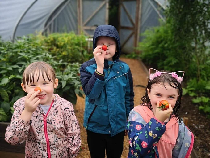 Children holding tomatoes in front of their noses whilst smiling and stood in a greenhouse. 