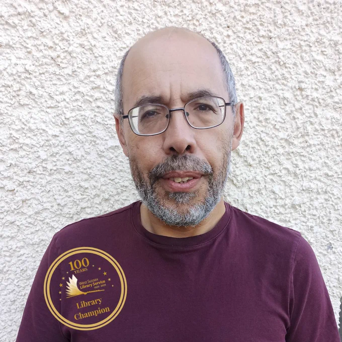 Digital volunteer Hatem in a burgundy t-shirt stood in front of a white brick wall with a golden library champion stamp on the picture.  