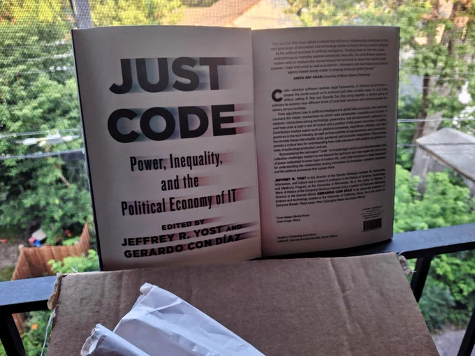 Box with two copies of JUST CODE adjacent to each other one showing front cover and one back cover on my balcony in my condo in Minneapolis with trees in the background.
