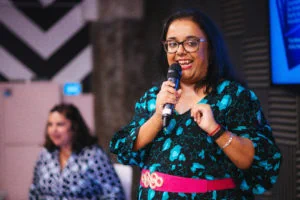 Advita with glasses and long black hair, wearing a patterned blue dress and a pink belt, stands confidently in front of a microphone. She is smiling and gesturing while speaking to an audience.