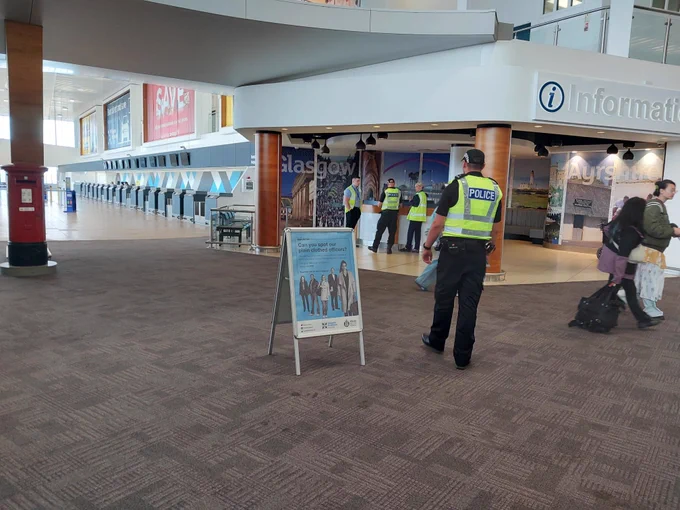 Photo shows uniformed police officers in an airport check in area. To the left of a uniformed officer is a poster A Frame. The frame has a poster with a blue background and photos of people in black and white.