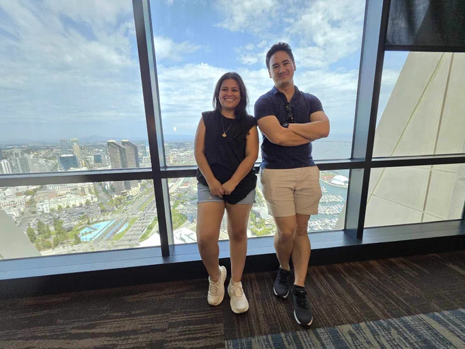 Janessa, wearing a dark, sleeveless shirt, light blue jean shorts and white sneakers and Oliver, wearing a navy polo, beige shorts, and black sneakers, standing in front of a glass window that overlooks downtown San Diego.