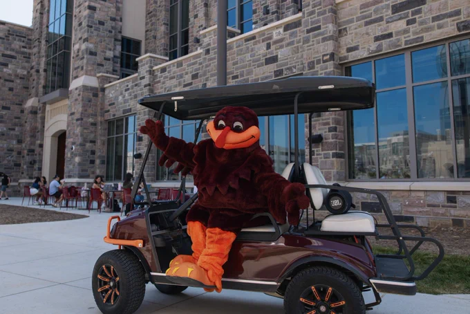 The HokieBird sits on a golf cart in front of Hitt Hall with his arms open wide. Students are seen in the background sitting at tables.