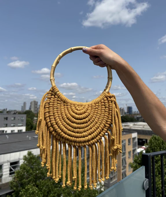 Yellow crescent moon shaped macramé bag with fringing around the edges and circular bamboo handles, held up over the rooftops of London on a summers day.