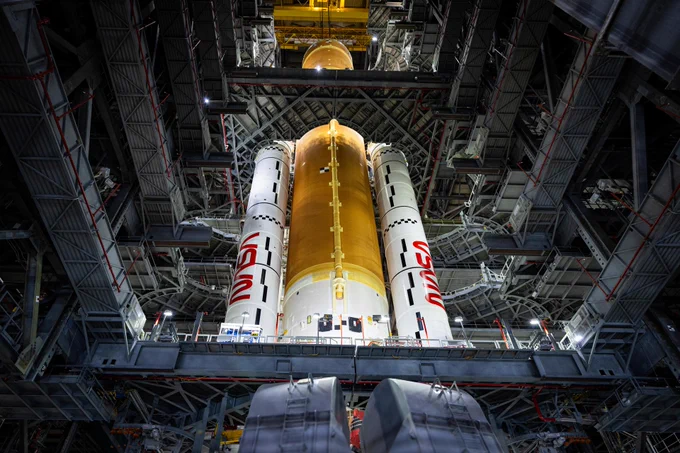 Teams with NASA’s Exploration Ground Systems and primary contractor Amentum, integrate the Space Launch System Moon rocket with the solid rocket boosters onto mobile launcher 1 inside High Bay 3 of the Vehicle Assembly Building at NASA’s Kennedy Space Center in Florida on Mar. 23, 2025. The core stage of the rocket has orange paneling and the booster on either side are white with the red NASA logo. The perspective of the image is from below the rocket looking up. Credit: NASA/Frank Michaux