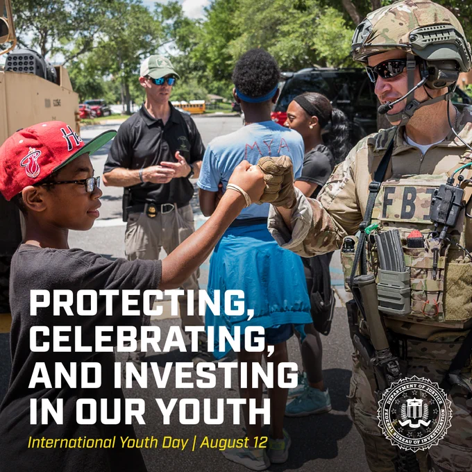 A young boy wearing a red cap fist bumps an FBI agent in tactical gear during a community event on a sunny day. The FBI seal is featured in the corner.