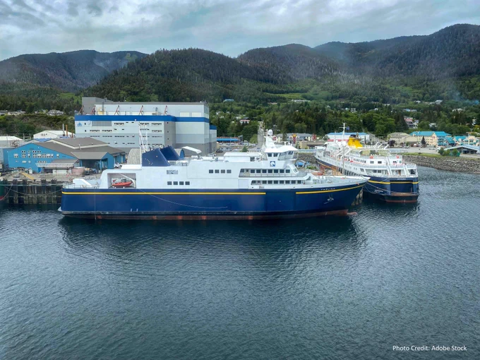 Two ferries docked at a port in a coastal town surrounded by forested hills. The ferry in the foreground is named Kennicott, and both are painted in blue and white. Cloudy skies and a calm sea add to the serene setting.
