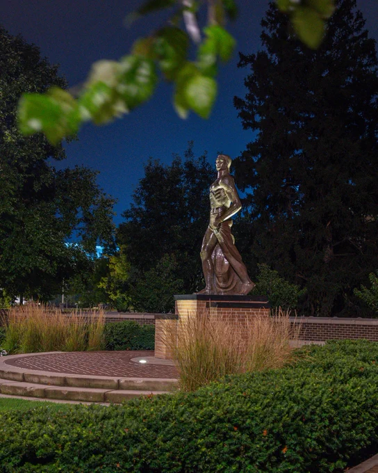 A bronze statue of the Michigan State University, Sparty stands illuminated at night on the Michigan State University campus.