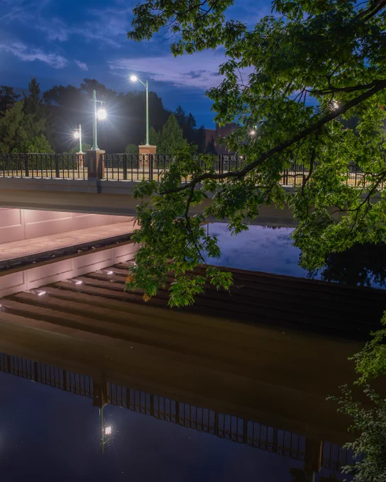 A bridge with illuminated railings crosses a river at night, reflecting in the calm water beneath.
