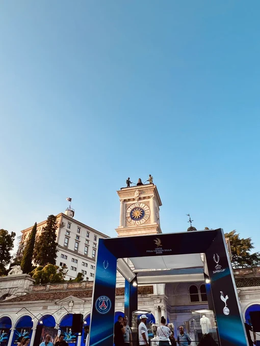 Picture from the UEFA Super Cup Fan Festival in the city centre of Udine. There is a dark blue arch way display the club badges of PSG and Tottenham with UEFA Super Cup Trophy displayed in the middle. The Loggia e Tempietto di San Giovanni is in the background.