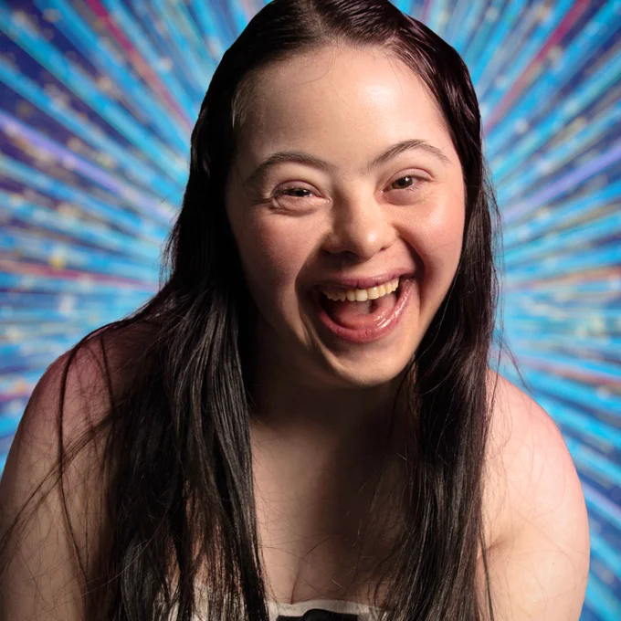A head and shoulders shot of Ellie Goldstein, a young woman with long brown hair, smiling to camera against a glittering blue background