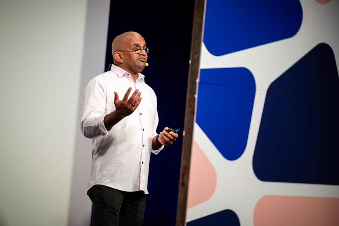 A bald man in a white shirt speaks expressively on stage, holding a small remote. He stands in front of a colorful backdrop with large abstract blue and pink shapes, wearing a headset microphone.