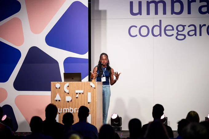 A woman with long braided hair, wearing glasses, a navy sleeveless top, and jeans, speaks passionately at a podium on the Umbraco Codegarden stage. She gestures with both hands while addressing the seated audience.