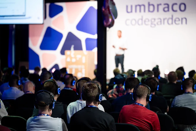 An audience at the Umbraco Codegarden conference wears wireless headphones while listening to a speaker on stage. The speaker appears blurred in the background, standing at a podium in front of a backdrop with the Umbraco logo and colorful abstract patterns.