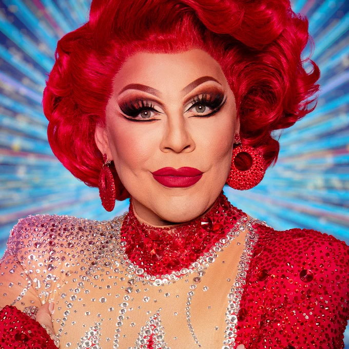 A head and shoulders shot of La Voix, a drag performer, wearing a red wig, glittering red dress, red earrings and a bold statement lip.