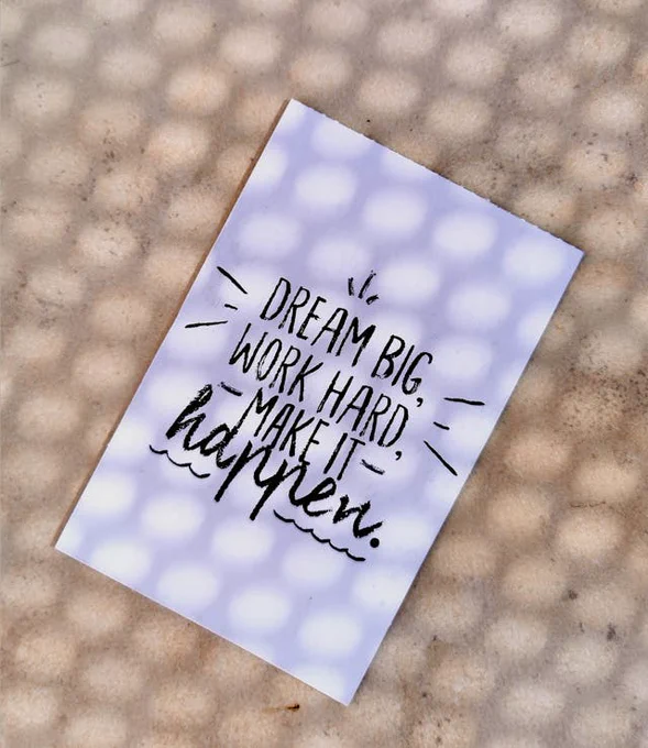 A motivational card with the text "DREAM BIG, WORK HARD, MAKE IT HAPPEN." It is placed on a textured surface that appears to be dotted or patterned.