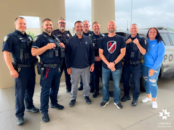 Group of eight Lincoln Police Department officers and one civilian smiling together at an outdoor event under a cloudy sky.