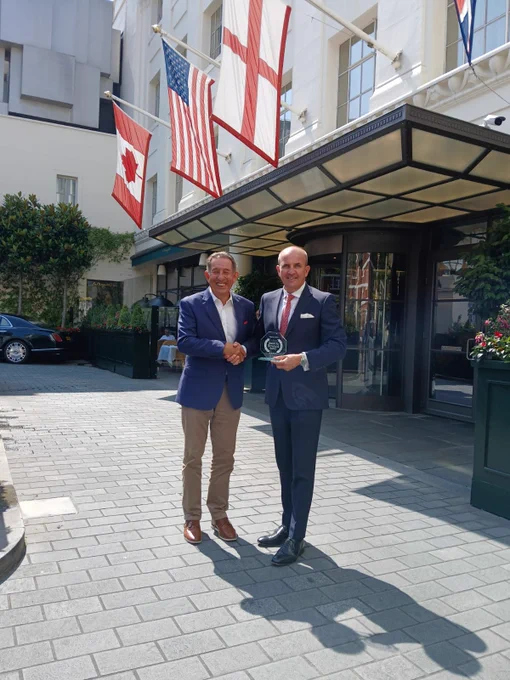 R2R Director Adam - in a navy blazer, white shirt and tan chinos - stands with Beaumont CEO Stuart Proctor - dressed in a navy suit, white shirt and red tie - outside the entrance to the hotel. The flags of Canada, USA and England (left to right) hang over the entrance. Stuart and Adam are shaking hands and Stuart is holding a glass R2R partner trophy.