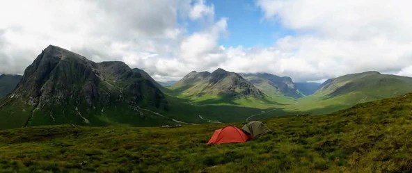 Photograph of an orange tent and a green tent are set up on the hillside, with four mountains behind them. Photo by the author.