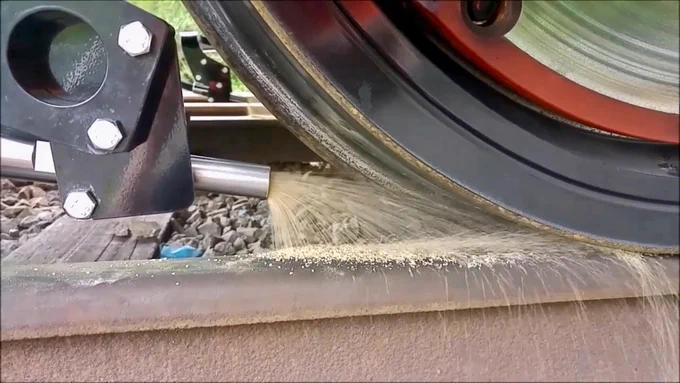 A close-up shows a train wheel on a track, with sand being dispensed between them. The wheel is black with a red accent. A metal nozzle directs the sand onto the rail. The track is light brown.