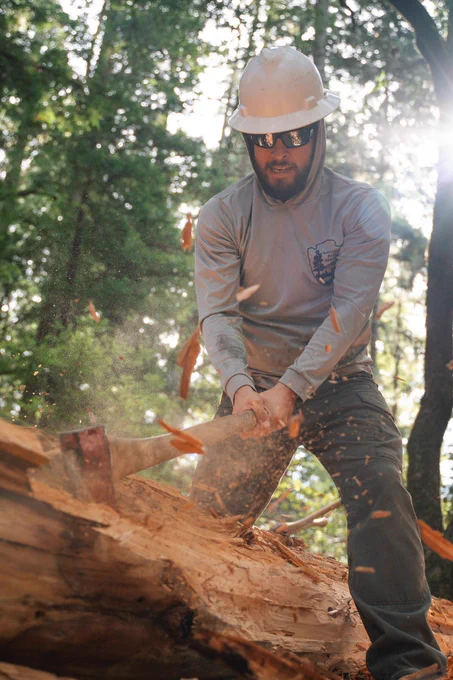 A person wearing a safety hat chops a log with an axe in the woods.