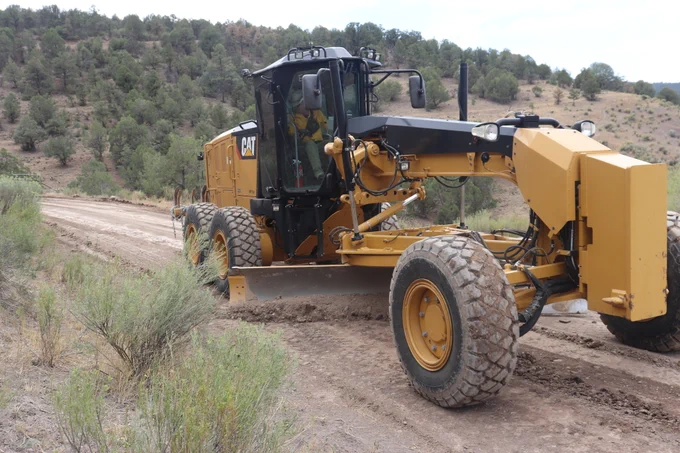 A graders uses its blade to smooth out ruts along a forest road.