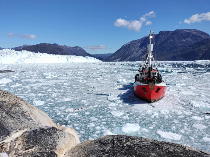 Frontal view of a boat from land with a glacier and a fjord environment in the background.