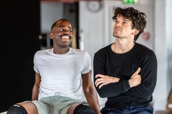 Ncuti Gatwa and Edward Bluemel in rehearsals for Born With Teeth, Gatwa smiles at Bluemel, who is crossed arm and looking contemplative