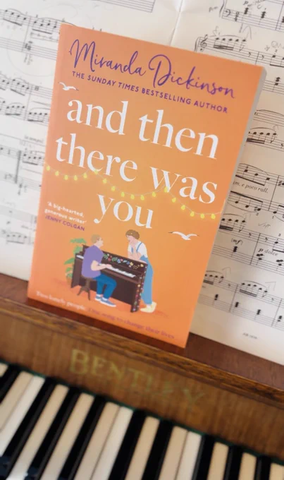 A paperback of my brand new book, And Then There Was You, resting on the music stand of my piano, with sheet music behind it. The cover shows a man sitting at a painted piano with a woman leaning against it, listening to him play. Strings of white lights loop above with two seagulls flying. The tagline reads: “Two lonely people. One song to change their lives.”